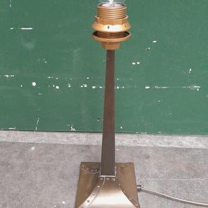 Puede incluir: Una lámpara de mesa de estilo vintage con una bombilla de vidrio transparente. La lámpara tiene una base cuadrada y un tallo delgado de metal marrón oscuro. El portalámparas y la base tienen un acabado dorado. La lámpara está enchufada.