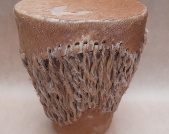 May include: A small, brown leather drum with a woven rope rim. The drum is made of natural materials and has a rustic appearance.