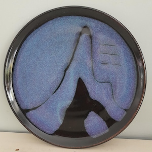 Può includere: Piatto rotondo in ceramica con bordo marrone scuro e design astratto blu e nero. Il centro presenta un motivo blu strutturato con accenti neri, creando un aspetto artistico unico. Il piatto è adatto per servire cibo o come elemento decorativo.