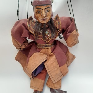 Peut inclure: Marionnette vintage avec une tête en bois sculpté et un costume en tissu marron et beige. La marionnette a de longs cheveux noirs et est ornée d'éléments décoratifs. Les ficelles sont visibles, indiquant son utilisation pour le spectacle.
