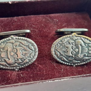May include: A pair of silver cufflinks with an ornate design. The cufflinks are oval shaped and have a detailed pattern of leaves and flowers. The cufflinks are in a velvet box.