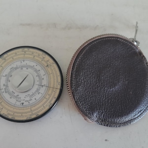 Może przedstawiać: Zabytkowe okrągłe suwmiarka z czarną obwódką i kremową tarczą, obok ciemnobrązowego skórzanego etui. Suwmiarka posiada różne skale i centralną tarczę. Etui jest okrągłe z zapięciem na zamek błyskawiczny.