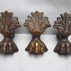 Puede incluir: Tres patas de muebles antiguas de color bronce con un diseño detallado de hojas. Cada pata tiene una base en forma de garra y una parte superior decorativa con patrones intrincados. Las patas son probablemente para un mueble antiguo.