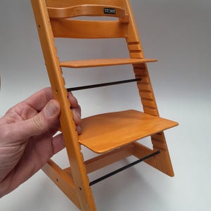 Puede incluir: Silla alta de madera naranja con asiento y reposapi&eacute;s ajustables. La silla alta tiene un respaldo curvo y una barra de metal negro para soporte. La marca "STOKKE" es visible en el respaldo.