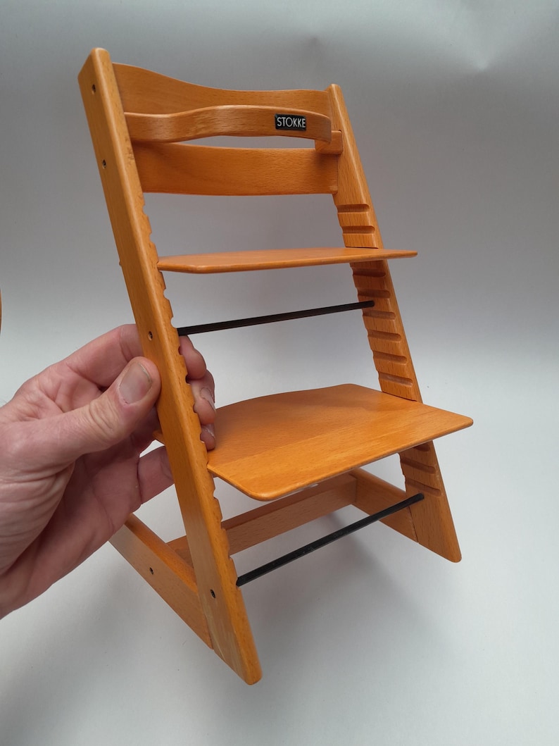 Puede incluir: Silla alta de madera naranja con asiento y reposapi&eacute;s ajustables. La silla alta tiene un respaldo curvo y una barra de metal negro para soporte. La marca "STOKKE" es visible en el respaldo.