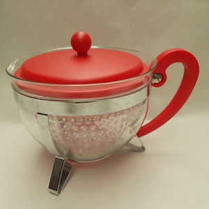 May include: A clear glass teapot with a red lid, handle, and knob. The teapot has a silver band around the middle and sits on three silver legs. The handle is curved and the lid is round.