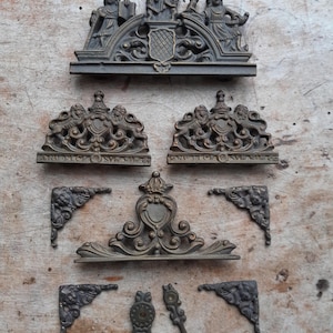 Può includere: Un assortimento di elementi decorativi antichi in bronzo. La collezione comprende stemmi ornati, staffe angolari e accenti decorativi. I pezzi presentano dettagliate incisioni a rilievo di figure, scudi e motivi floreali. L'hardware è disposto su una superficie di legno stagionato.