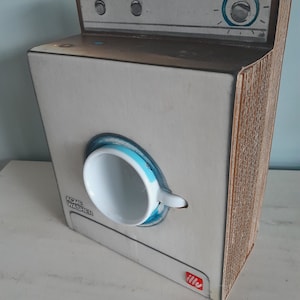 Op de afbeelding: Een wit kartonnen model van een wasmachine met een blauw-witte koffiekop in de opening. De wasmachine heeft een bruine bovenkant met een knop en twee knoppen. De tekst "ACME WASHER" staat gedrukt op de zijkant van de machine. De onderkant van de machine heeft een rood "illy"-logo.