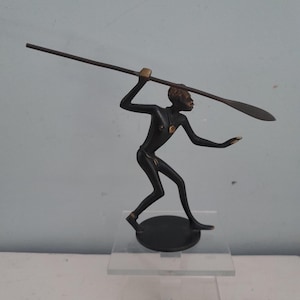 Könnte beinhalten: Eine dunkel bronzefarbene Metallfigur, die eine Person in einer dynamischen, athletischen Haltung darstellt, die einen langen, schlanken Speer horizontal über dem Kopf hält. Die Figur ist auf einem runden Sockel montiert.
