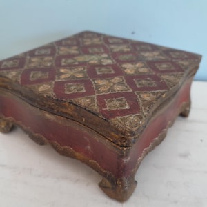 Puede incluir: Una pequeña caja de madera roja con un patrón geométrico decorativo pintado en oro en la tapa. La caja tiene cuatro patas y está hecha de madera.