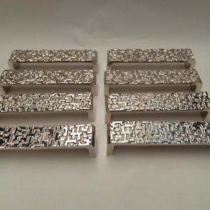 Könnte beinhalten: Acht silberfarbene Schrankgriffe mit einem strukturierten, geometrischen Design. Jeder Griff hat eine rechteckige Form mit einem gebogenen Griff und einem detaillierten Muster auf der Vorderseite. Die Griffe sind in zwei Reihen zu je vier Stück angeordnet.