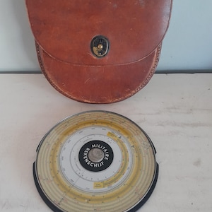 Może przedstawiać: Zabytkowe brązowe skórzane etui z metalowym zapięciem i okrągłym suwakiem logarytmicznym z napisem "MILITAIRE REKENSCHIJF". Suwak logarytmiczny ma żółte i białe oznaczenia.