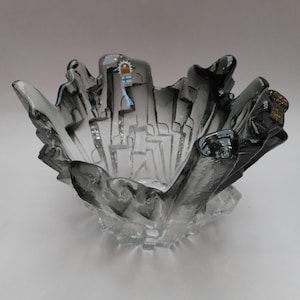 May include: A clear glass bowl with a unique, textured design. The bowl has a dark gray rim and a textured, irregular shape. The interior features a cityscape design. A small sticker with a flag is visible on the rim.