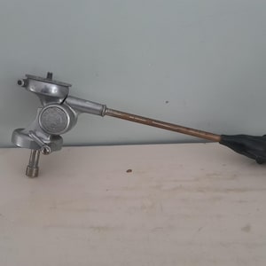 Può includere: Un dispositivo meccanico color argento con un lungo braccio color bronzo e un'impugnatura nera. Il dispositivo ha un emblema circolare e componenti regolabili. Probabilmente uno strumento o parte di una macchina più grande.