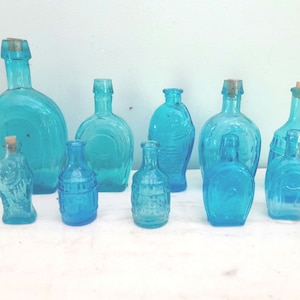 Op de afbeelding: Collectie van negen blauwe glazen flessen met kurken stoppen. De flessen zijn in verschillende vormen en maten, sommige met gegraveerde ontwerpen.
