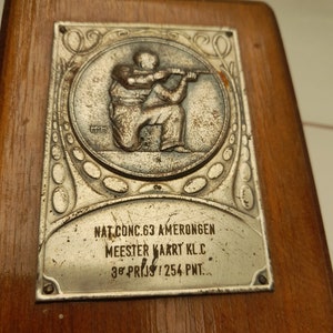 Puede incluir: Una placa de madera con una placa de metal plateado. La placa presenta una escultura en relieve de una persona disparando un rifle. El texto en la placa dice "NAT. CONC. 63 AMERONGEN MEESTER KAART K.L.C. 3e PRIJS 254 PNT."