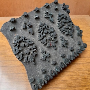 May include: A dark, weathered wooden block used for textile printing, featuring raised, intricate floral and leaf patterns carved into the surface. The block has an irregular, roughly rectangular shape with visible signs of age and use.