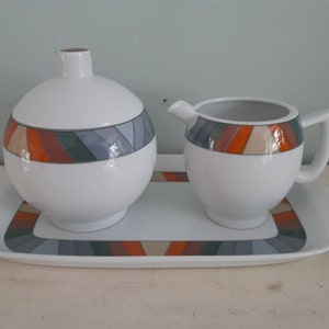 Op de afbeelding: Een witte keramische suikerpot met deksel, een melkkan en een serveerschaal. Elk stuk heeft een geometrisch ontwerp van oranje, grijze en groene strepen. De set is ontworpen voor het serveren van koffie of thee.
