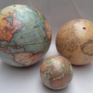 Op de afbeelding: Drie globes in antieke stijl van verschillende formaten. De grootste globe heeft een blauw en bruin kleurenschema met gedetailleerde cartografie. De middelste globe is bruin met ingewikkelde kaartdetails. De kleinste globe heeft een veelkleurig ontwerp.