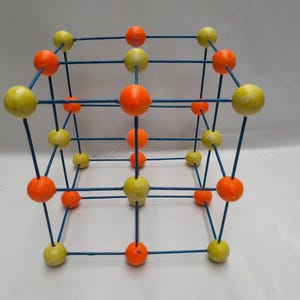 Könnte beinhalten: Ein dreidimensionales kubisches Molekülmodell mit blauen Verbindungsstäben und abwechselnd orangefarbenen und blassgelben Kugeln, die in einem Gittermuster angeordnet sind, um atomare Bindungen und Positionen darzustellen.