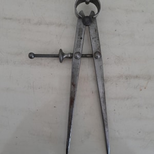 Puede incluir: Un compás de puntas de metal vintage con dos patas afiladas conectadas por una bisagra circular y un tornillo de ajuste roscado. La superficie metálica muestra signos de desgaste y oxidación. El texto Moore & Wright Sheffield England está grabado.