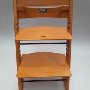 Puede incluir: Silla alta de madera de color marr&oacute;n claro con asiento y reposapi&eacute;s ajustables. La silla tiene un respaldo curvo y una barra de metal negra. La marca "STOKKE" es visible en el respaldo. Dise&ntilde;ada para ni&ntilde;os.