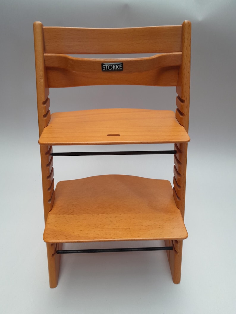 Puede incluir: Silla alta de madera de color marr&oacute;n claro con asiento y reposapi&eacute;s ajustables. La silla tiene un respaldo curvo y una barra de metal negra. La marca "STOKKE" es visible en el respaldo. Dise&ntilde;ada para ni&ntilde;os.