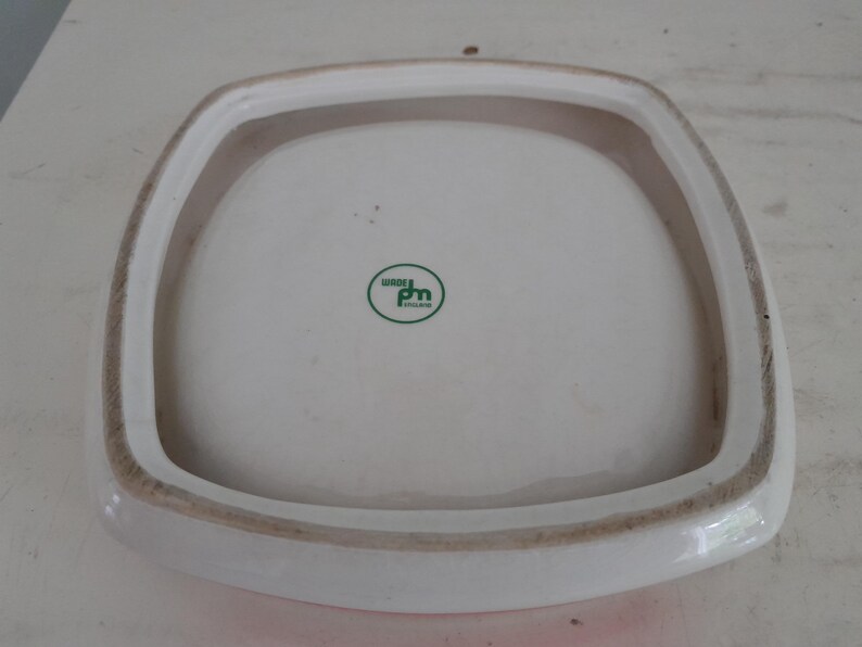 May include: A square, white ceramic plate with a raised, rounded rim. The rim is bordered by a brown band. The plate has a green and white logo that reads "Wade England".