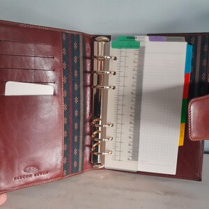 May include: Open brown leather planner with a gold-colored ring binder. The planner has a card slot and a tabbed section with lined and graph paper. The planner is open, revealing the interior.