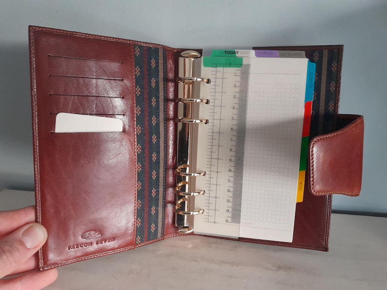 May include: Open brown leather planner with a gold-colored ring binder. The planner has a card slot and a tabbed section with lined and graph paper. The planner is open, revealing the interior.