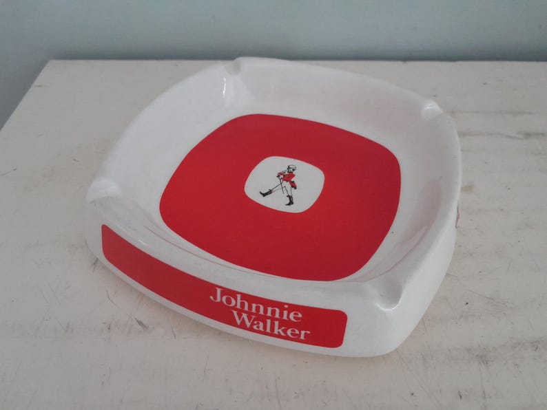 May include: White ashtray with a red center and a red rectangular label that reads "Johnnie Walker". The center features a small white square with a figure. The ashtray has a curved shape and is designed for cigarettes.