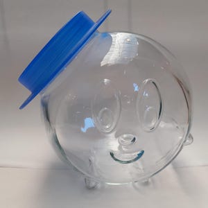 Op de afbeelding: Een glazen pot met een glimlachend gezicht en drie poten. De pot heeft een blauw plastic deksel.