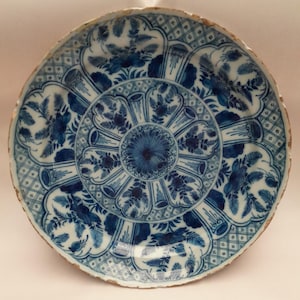 Puede incluir: Un plato de cerámica redondo y antiguo con un diseño floral azul y blanco. El plato presenta un motivo floral central rodeado de patrones florales y de hojas estilizados. El borde tiene un patrón de diamantes y una apariencia envejecida y desgastada.
