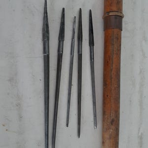 Puede incluir: Un conjunto de punzones metálicos de varios tamaños, con un tubo de almacenamiento de madera. Los punzones son plateados y tienen puntas afiladas. El tubo de madera es cilíndrico, con una banda metálica oscura. Herramientas sobre una superficie clara.