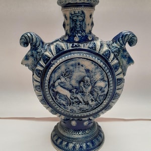 Może przedstawiać: Niebiesko-biały ceramiczny wazon o okrągłym korpusie i trzech uchwytach w kształcie głowy barana. Wazon zdobi płaskorzeźba przedstawiająca mężczyznę polującego na jelenia w lesie.