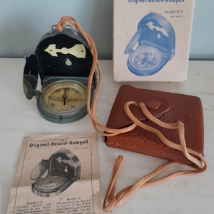 May include: An open, vintage Bézard-Kompaß compass with a mirror, a leather case, and a box. The compass is olive green with a cream-colored directional arrow. The box and paperwork are visible, with text in German.