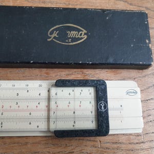 May include: A vintage slide rule and case. The slide rule is off-white with black and red markings, and a clear plastic cursor. The case is black with gold lettering that reads "Gamma".