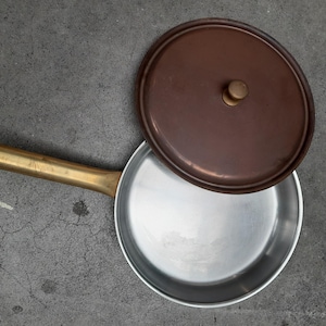 Puede incluir: Una pequeña cacerola redonda de color plateado con un mango largo de color latón. La tapa marrón está ligeramente entreabierta, revelando el interior de la cacerola. El mango tiene un agujero para colgar.