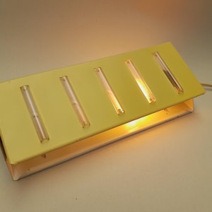 May include: A vintage yellow wall-mounted light fixture with five rectangular clear plastic light diffusers. The fixture is rectangular and has a metal frame. The light is on, illuminating the diffusers.