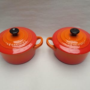 May include: Two round enamelled cast iron Dutch ovens by Le Creuset, featuring a vibrant gradient of orange and red. Each pot includes two side handles and a matching lid with a black knob. The brand name Le Creuset is embossed on the lids.