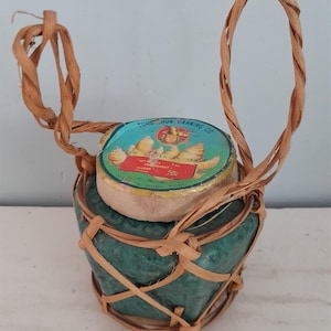 Op de afbeelding: Een kleine, groene keramische pot met een geweven handvat en rand, met een blikje stemgember in siroop. Het blikje is turquoise met gouden en rode tekst, en de pot is omwikkeld met geweven stro.