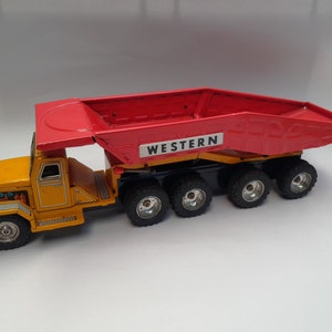 Peut inclure: Un camion à benne basculante jouet jaune avec une benne rouge à charnières qui dit "WESTERN" sur le côté. Le camion a quatre roues noires et une cabine détaillée.