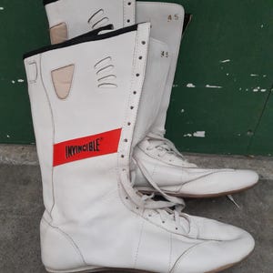 Könnte beinhalten: Paar weiße Lederstiefel mit Schnürsenkeln, mit einem roten Streifen mit dem Wort "INVINCIBLE". Die Stiefel haben ein hoch geschnittenes Design mit schwarzem Besatz und der Zahl "45" auf der Seite. Sportschuhe.