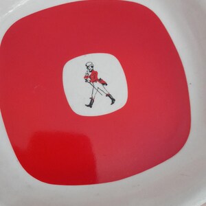 May include: White, square-shaped serving tray with a red center. The center features a white square with a drawing of a man in a red coat and black boots. The tray has a glossy finish.