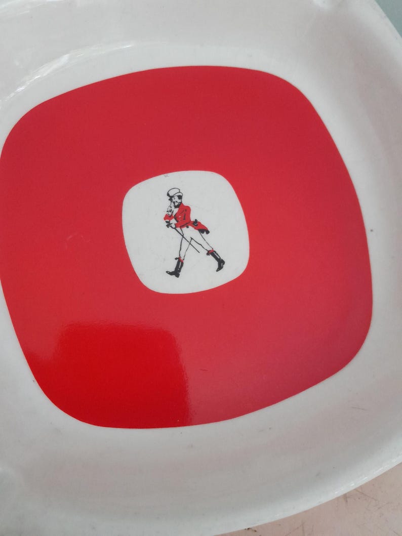May include: White, square-shaped serving tray with a red center. The center features a white square with a drawing of a man in a red coat and black boots. The tray has a glossy finish.