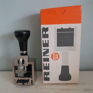 Puede incluir: Un sello metálico y negro con un mango negro junto a una caja blanca y naranja. La caja dice "REINER" en letras negras grandes y "REINER COLOR BOX" en letras más pequeñas. El sello es un dispositivo manual.