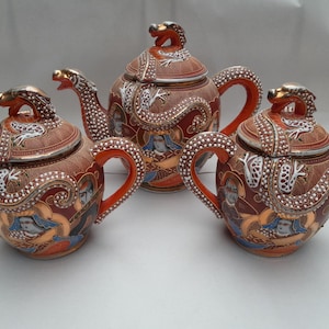 Peut inclure: Ensemble de trois théières japonaises antiques. Les théières sont marron avec des poignées et des accents orange, avec des becs et des couvercles en forme de dragon. Les théières sont décorées d'œuvres d'art détaillées et de garnitures dorées.