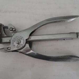 May include: A vintage silver-colored Simplex stapling plier. The tool has a long, cylindrical body with curved handles and a stapling head. The text "SIMPLEX STAPLING PLIER" is visible on the tool. The tool is designed for stapling.