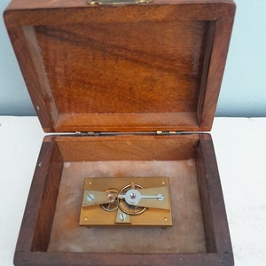 Könnte beinhalten: Ein kleiner, komplizierter Uhrwerkmechanismus wird in einer Holzkiste mit einem Messingverschluss gezeigt. Der Mechanismus ist aus Messing gefertigt und hat ein Rubin-Juwelenlager. Die Kiste ist mit braunem Filz ausgekleidet.