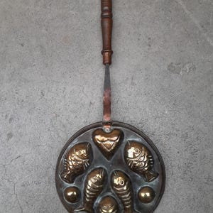 Op de afbeelding: Antieke bronzen visvormige pan met een houten handvat. De pan heeft hart- en visontwerpen, met een lange handgreep. Een uniek keukengerei.